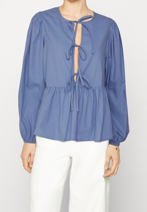 Blauwe blouse met lange mouwen, drie strikjes aan de voorkant en een geplooide taille, gecombineerd met een witte broek bij een staand persoon.