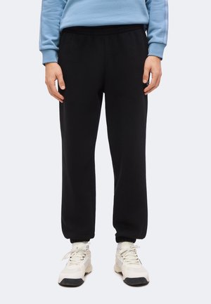 Personne portant un pantalon de jogging noir, des baskets blanches et un haut à manches longues bleu clair sur un fond uni.