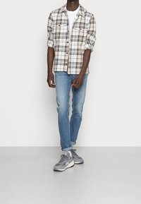 Camisa de cuadros en beige, gris y blanco; combinada con jeans de mezclilla claros y zapatillas grises. Presenta detalles de bolsillos con botones y mangas enrolladas.