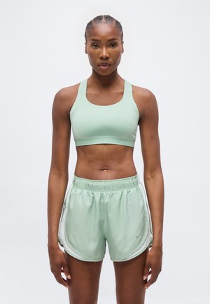 Atletische vrouw staand, draagt een lichtgroene sportbeha en bijpassende shorts, kijkt recht vooruit met een neutrale uitdrukking tegen een effen achtergrond.