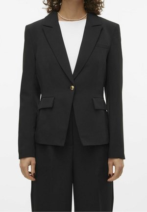Kvinde iført en sort skræddersyet blazer med en guldknap over en hvid top og sorte bukser, accessoriseret med en perle halskæde.