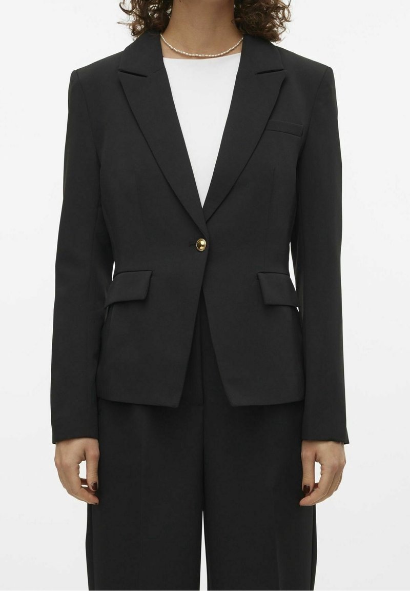 Femme portant un blazer noir taillé avec un bouton doré sur un haut blanc et un pantalon noir, accessoirisée d'un collier de perles.