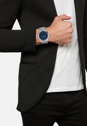 Sølvfarvet ur med en minimalistisk blå urskive, rem i rustfrit stål og glat kasse. Båret på et håndled, parret med en sort blazer.