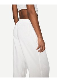 Person i vita linnebyxor med vida ben och en kortärmad topp, som visar midjan och höger arm mot en enkel vit bakgrund.