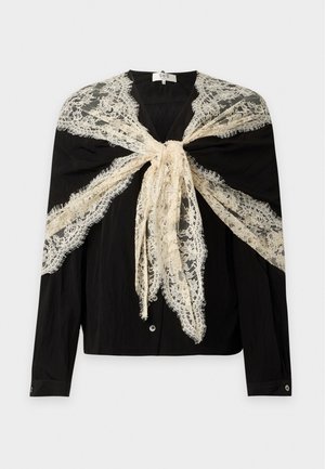 Femme aux longs cheveux foncés portant une tenue noire ample et un grand châle en dentelle beige noué en nœud papillon sur ses épaules.