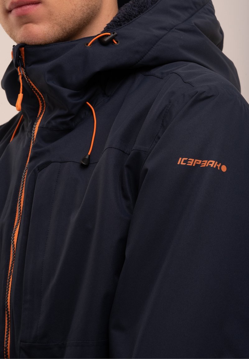 Homme portant une veste à capuche bleu marine avec une fermeture éclair et des cordons orange, avec le logo "ICEPEAK" sur le haut du bras.