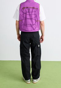 Fioletowa lekka kamizelka z suwakiem z przodu, regulowanymi paskami i siateczkową fakturą; w komplecie z czarnymi spodniami cargo i srebrnymi tenisówkami.