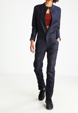 Woman walking wearing a dark navy jumpsuit with a partially unzipped front and a rust-colored cropped top underneath, paired with black sneakers.