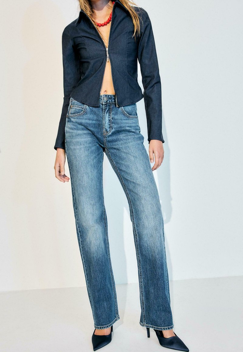 Femme portant une veste zippée moulante de couleur foncée, un collier de perles rouges, un jean bleu délavé droit, et des talons hauts pointus noirs, debout sur un sol blanc.