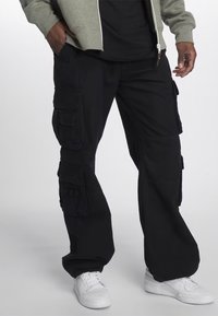 Černé cargo kalhoty vyrobené z odolného materiálu, s několika kapsami, volným střihem a mírným leskem. V kombinaci s bílými teniskami.