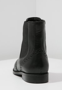 Botas de cuero negro hasta el tobillo con paneles elásticos en los laterales, detalles de brogue, superficie lisa y un tacón bajo apilado. Vista trasera que muestra los detalles de las costuras.