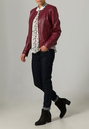 Vrouw draagt een bordeauxrood leren jack, een witte blouse met patroon, donkere skinny jeans met omslagen en zwarte enkellaarsjes, staande voor een grijze achtergrond.