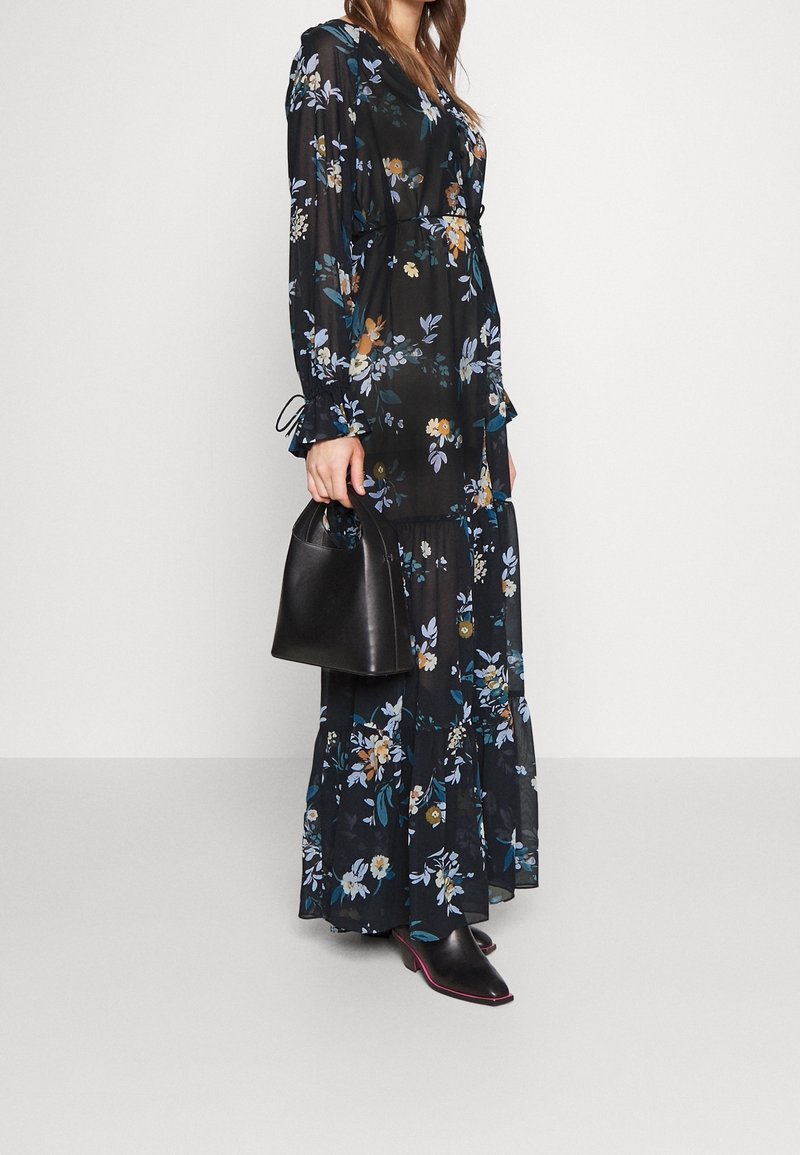 Femme portant une longue robe noire à fleurs tenant un sac à main noir, associée à des chaussures pointues noires avec des semelles roses.