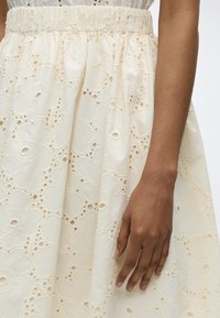 Το δεξί χέρι ακουμπά δίπλα σε μια φούστα σε εκρού χρώμα με κεντήματα με λουλούδια και ελαστική μέση.
