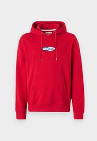Red cotton hoodie with a front pocket, drawstring hood, and a blue and white "GUESS JEANS" logo patch on the chest. Soft texture.