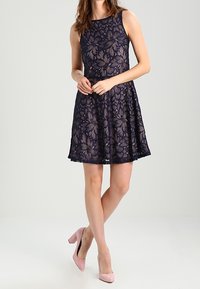 Merino de renda azul-marinho com corpete ajustado, decote redondo e saia rodada. Apresenta delicados padrões de folhas e é combinado com saltos altos rosa.
