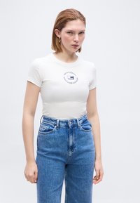 Νεαρή γυναίκα που φοράει λευκό εφαρμοστό κοντομάνικο μπλουζάκι με κυκλικό λογότυπο και ψηλόμεσο μπλε τζιν, μπροστά σε απλό ανοιχτόχρωμο φόντο.