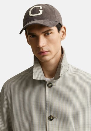 Jonge man met lichte huid die een geruit jasje met hoge kraag draagt en een donkere pet met een grote witte "G" aan de voorkant, neutrale achtergrond.