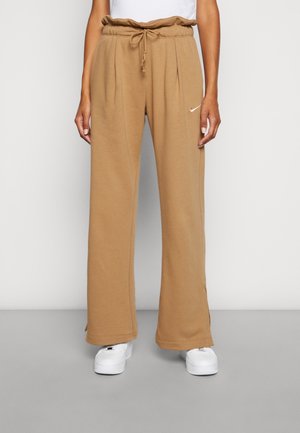 Kvinde iført camelfarvede, vidde sweatpants med elastisk talje og snor, parret med hvide sneakers, stående mod en ensfarvet baggrund.