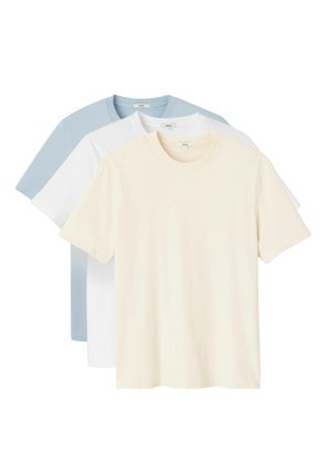 Jeune homme aux cheveux bruns mi-longs portant un t-shirt beige clair uni à manches courtes et un jean bleu, regardant vers sa gauche.