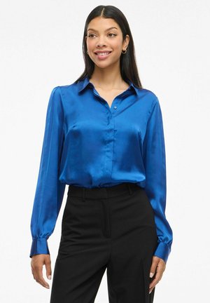 Satin blue blouse with long sleeves, button-down collar, and cuffs; paired with black tailored trousers. Smooth texture with a relaxed fit.