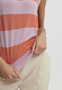 Suéter sin mangas a rayas en naranja y púrpura claro, con un dobladillo acanalado. Combinado con pantalones beige, exhibiendo un look casual.