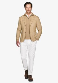Beige blazer med to forlommer over en cremet striktrøje, parret med hvide bukser og brune snørebåndsstøvler. Person går fremad.