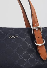 Laadukkaita tummansinisiä nylontaskuja, joissa on teksturoitu geometrinen kuvio, ruskeat nahkalenkit ja hopeinen solki. Brändätty "JOOP!" metallinvärisellä tekstillä.