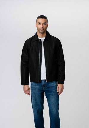 Mand med kort hår iført en sort lynlåstrøje over en hvid skjorte og blå jeans, stående mod en ensartet lys baggrund.