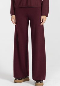 Kobieta ubrana w szerokie burgundowe dzianinowe spodnie, zestawione z dopasowanym długim rękawem oraz brązowymi butami na platformie, na białym tle.