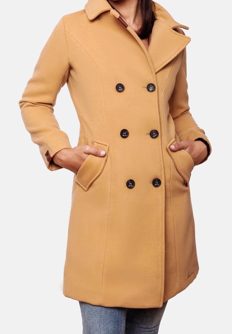 Femme portant un manteau croisé couleur camel avec des boutons noirs, un col, et les mains dans les poches avant, associé à un jean bleu.