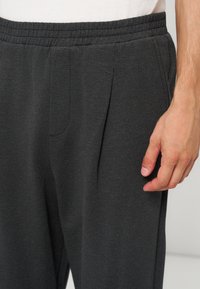 Gros plan sur un pantalon gris foncé à taille élastique avec des plis et une main reposant le long de la cuisse droite.