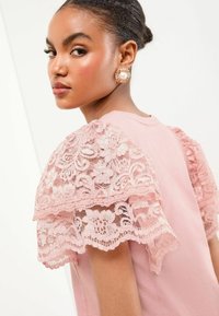 Haut rose blush avec des manches en dentelle florale, présentant des motifs complexes et des bords festonnés, fabriqué en tissu doux pour un aspect texturé.