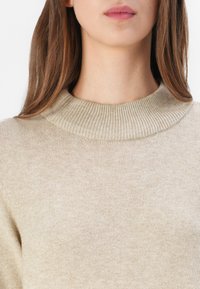 Maglione beige a maglia con colletto alto a coste e una texture liscia. Il tessuto appare leggero e leggermente elastico.