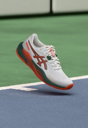 Chaussure de sport blanche et orange avec des accents verts planant au-dessus de la surface d'un court de tennis, près de la ligne de fond.