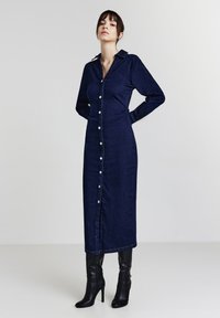 Robe chemise en denim bleu foncé avec des manches longues, des boutons sur le devant, un col et des fronces sur les côtés. Portée avec des bottes noires à talons jusqu'aux genoux.