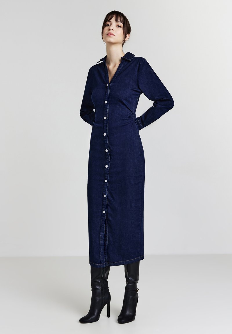 Robe chemise en denim bleu foncé avec des manches longues, des boutons sur le devant, un col et des fronces sur les côtés. Portée avec des bottes noires à talons jusqu'aux genoux.