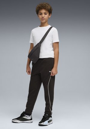 Niño con camiseta blanca, pantalones deportivos negros con una raya blanca, bolso cruzado negro y zapatillas negras y blancas, de pie frente a un fondo gris.