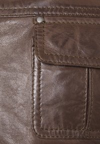 Bruin leermateriaal met een gestructureerd oppervlak, een rechthoekig zakje, metalen drukknop en contrasterende stiksel details langs de randen.