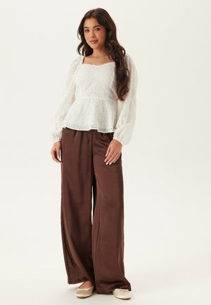 Kvinde med langt mørkt hår iført en hvid bluse med hulmønster og lange ærmer, brune bukser med vidde i benene og beige flade sko, stående mod en lys baggrund.