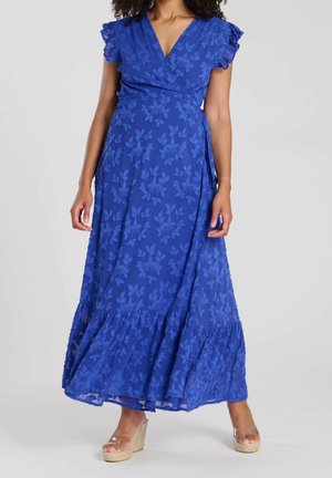 Femme portant une robe portefeuille fleurie bleue avec manches courtes volantées et ourlet, associée à des sandales compensées transparentes, debout sur fond blanc.