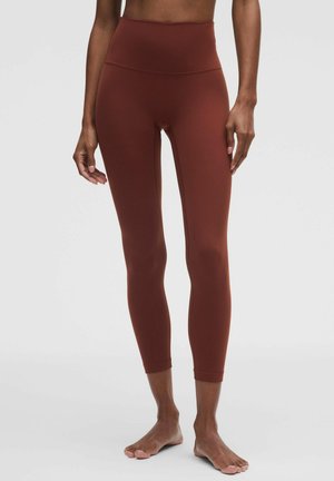 Donna che indossa leggings a vita alta color ruggine, in piedi a piedi nudi su uno sfondo chiaro e semplice.