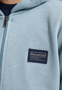 Ljusblå zip-up hoodie tillverkad av mjukt tyg, med en marinblå etikett som har "Petrol Ind" och text. Inkluderar en huva och en texturerad yta.