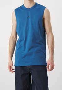 Niebieska bawełniana koszula bez rękawów z okrągłym dekoltem i małym czarnym logo na piersi. W komplecie z ciemnymi dżinsami.