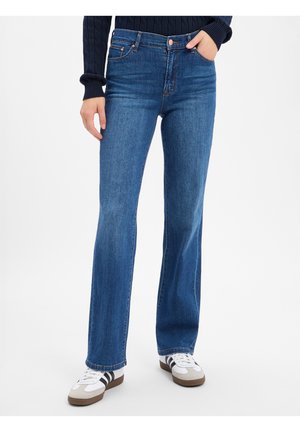 Persoon draagt blauwe jeans met rechte pijpen, een donkere trui en witte sneakers met zwarte strepen, staand tegen een witte achtergrond.