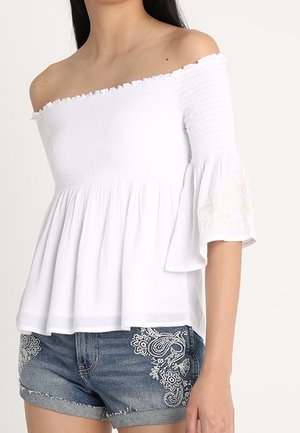 Femme portant un top blanc épaules dénudées avec des manches évasées et un short en jean avec une broderie florale blanche sur les poches avant.