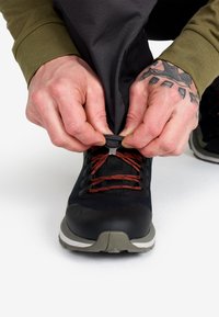 Händer med en tatuering justerar en svart byxled över en svart sko med röda snören på en vit yta.