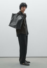 Sac fourre-tout en cuir noir avec une forme structurée, doté de deux poignées supérieures et de subtils détails de couture. Le modèle porte une tenue sombre avec des sandales.
