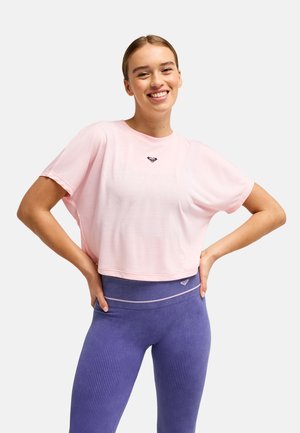 Femme souriante aux cheveux attachés porte un t-shirt court rose clair et des leggings violets côtelés taille haute, les mains sur les hanches.