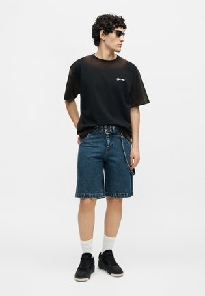 Junger Mann mit lockigem Haar, der schwarze Sonnenbrillen, ein schwarzes T-Shirt, Jeans-Shorts mit Gürtelkette, weiße Socken und schwarze Sneaker trägt.